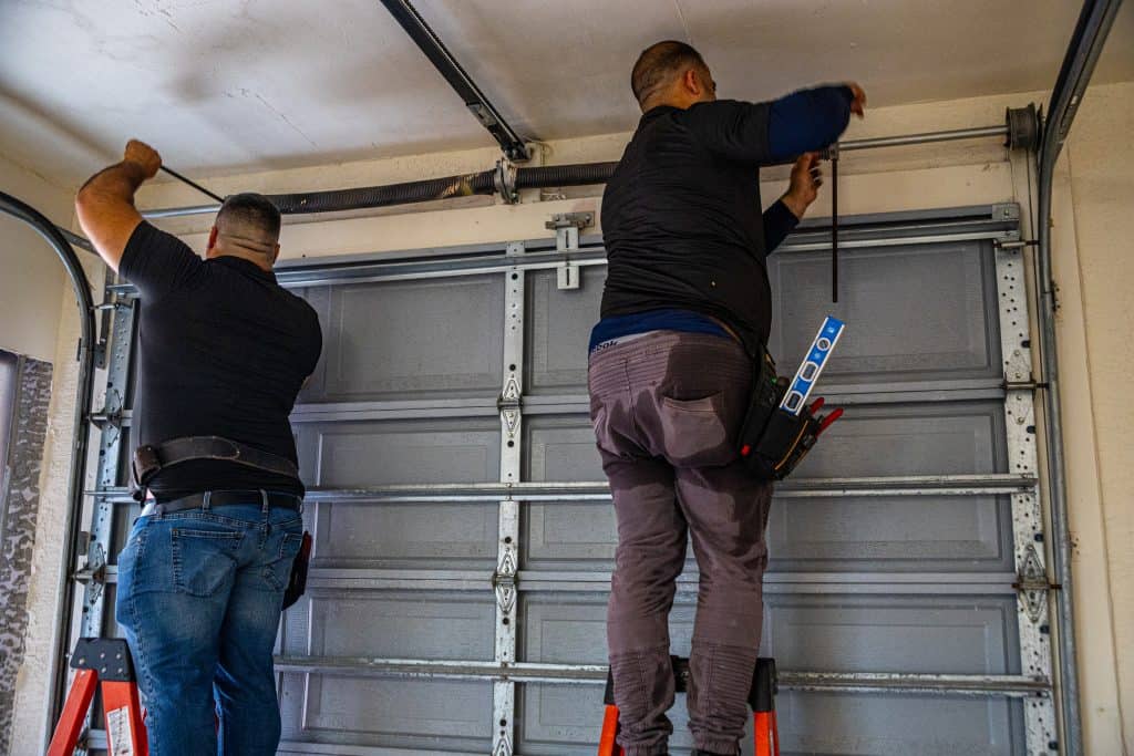 Two men standing on ladders, working together on a HOG TOWN Garage Door Repair, fixing or installing a metal garage door mechanism in a residential garage.