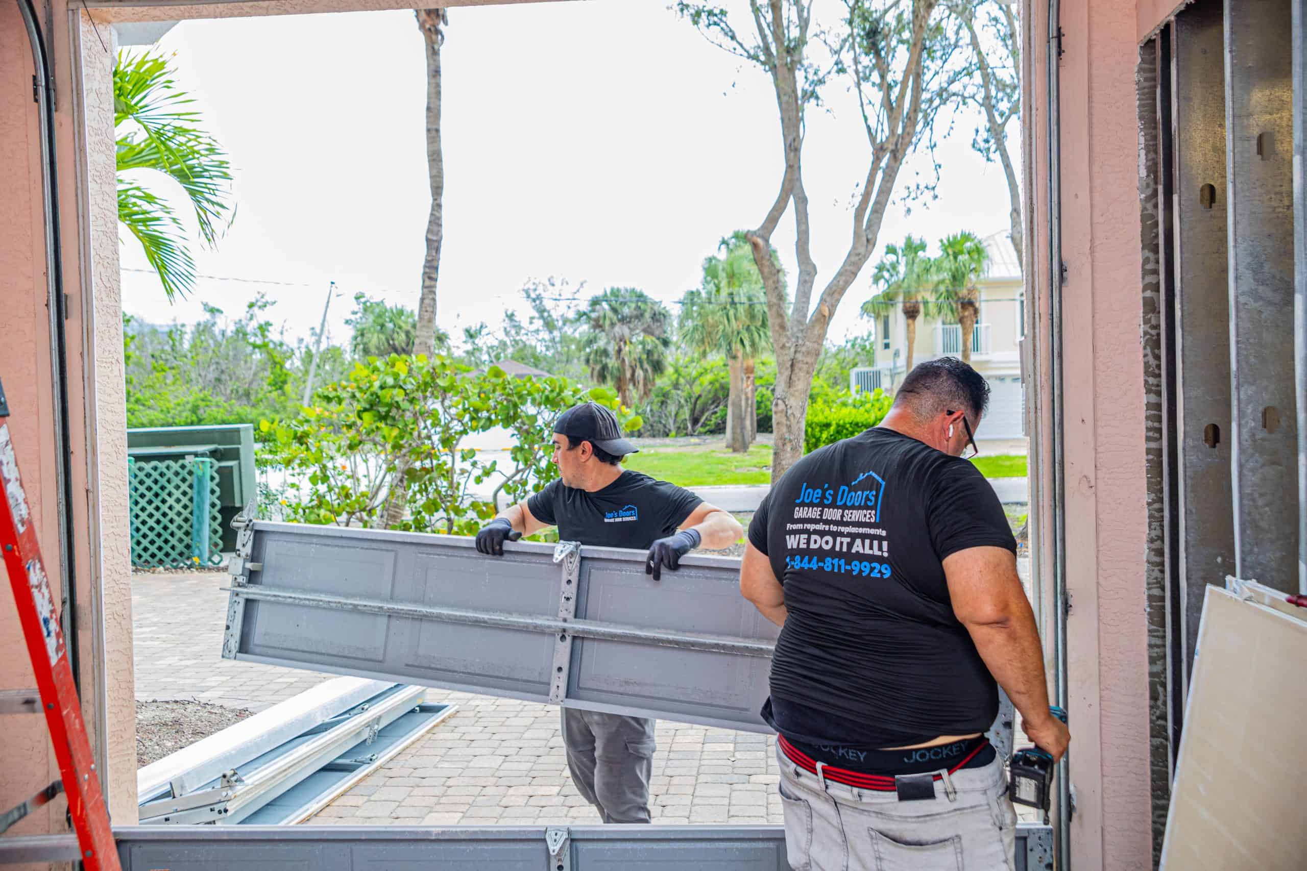 Two men from HOG TOWN Garage Door Repair are installing a garage door panel in a residential garage. One holds the door upright while the other guides it into place. Trees and houses are visible outside; both wear shirts with the company logo.