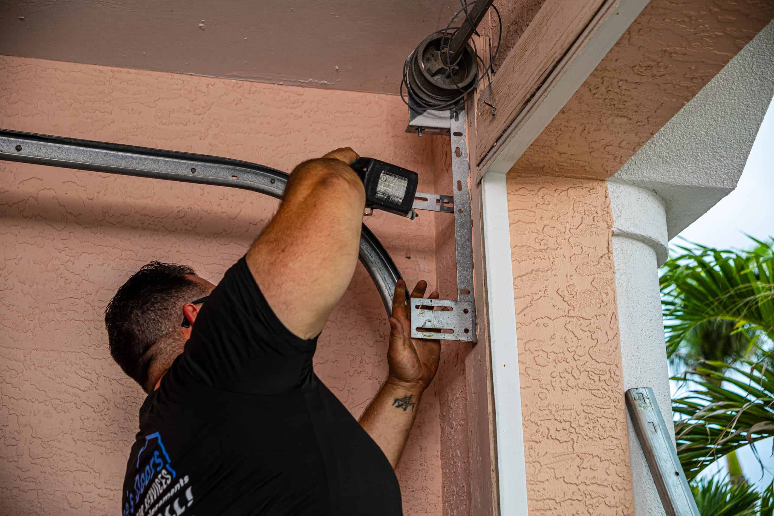 A person uses a power drill for Garage Door Repair, installing or fixing a metal garage door track on a peach-colored wall, with part of a white column and a palm tree visible outside—service by HOG TOWN.
