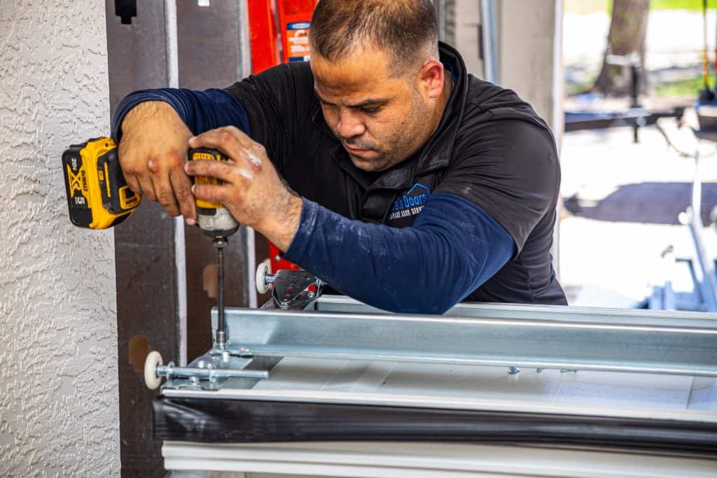 A person is using a power drill to install or repair a metal component, possibly part of a garage door. They are focused on their task, wearing a dark shirt and working near a textured wall. A red ladder is visible in the background.
