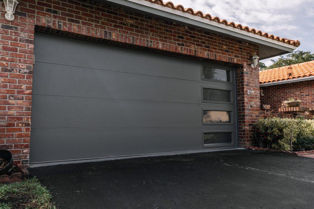 A modern garage door with horizontal panels and glass sections on the right side is surrounded by a brick facade, provided by a top garage door company in Broward County, FL. The driveway is paved with asphalt, accompanied by plants to the right of the door under a partly cloudy sky.