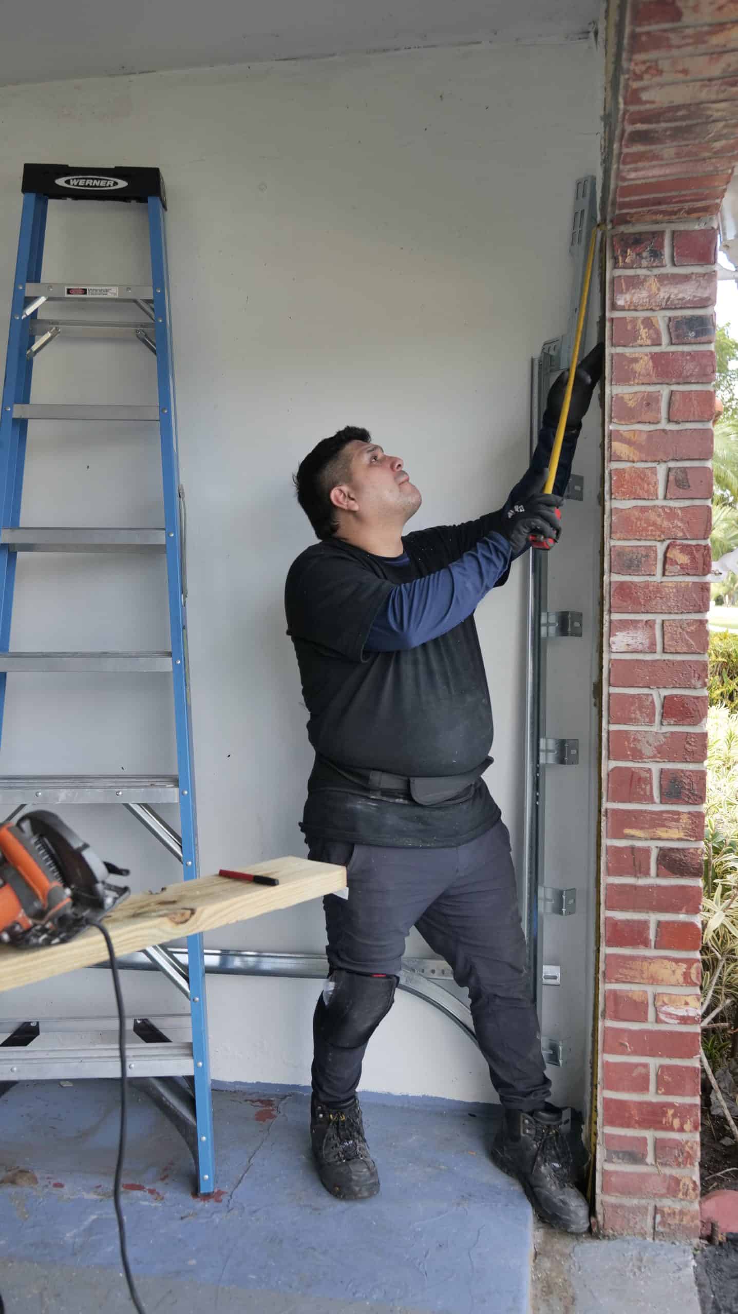 A person in work clothes and gloves uses a tape measure to check the height of a brick doorway. Nearby, a metal step ladder and workbench with tools hint at ongoing HOG TOWN Garage Door Services in the partially finished room.