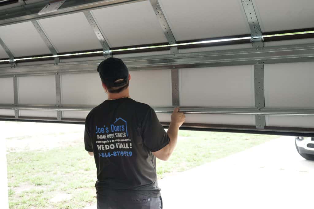 A person wearing a "Joe's Doors Garage Door Services" t-shirt is lifting a partially open garage door from inside, sunlight streaming in and a parked car outside—showcasing the work of this expert garage door company.