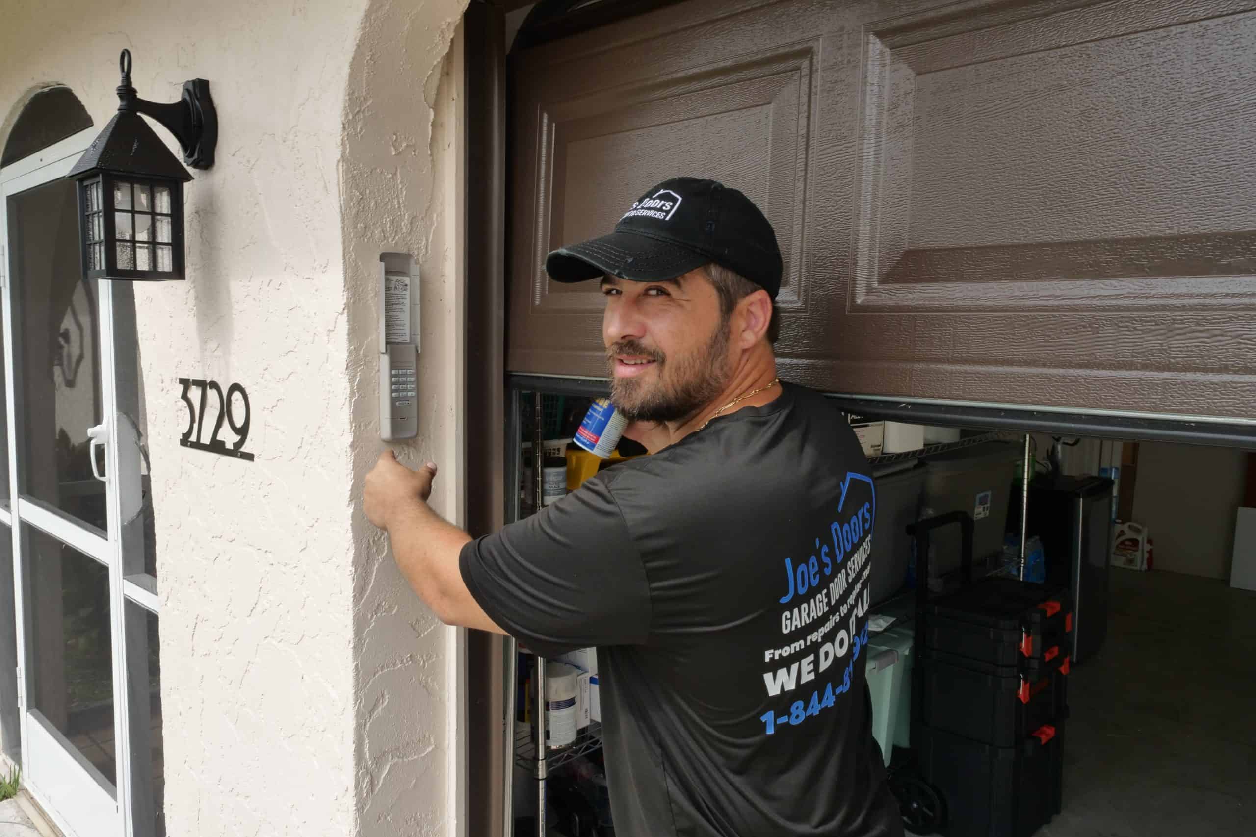 A man wearing a black cap and shirt stands by a partially open garage door, performing Garage Door Repair next to the entrance. He is smiling while holding tools, with a toolbox visible inside the HOG TOWN garage.