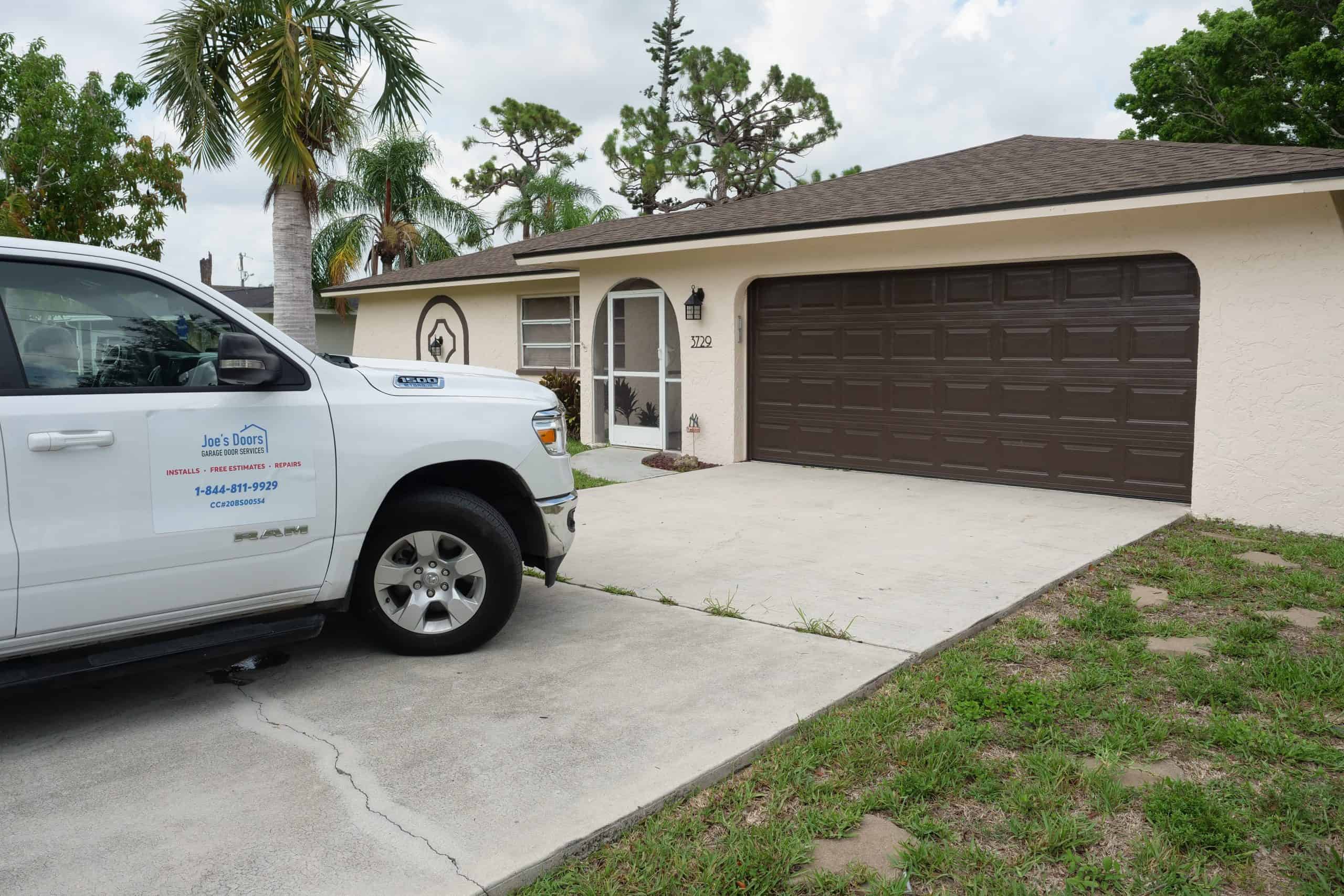A white pickup truck with a "Joe’s Doors" sign, advertising Garage Door Repair, is parked in the driveway of a beige single-story house in HOG TOWN, with palm trees and greenery all around.