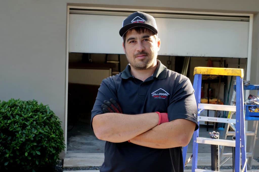 A man in a black polo shirt and cap stands with arms crossed, wearing work gloves, in front of a partially open garage. A blue ladder and some tools for garage door replacement are visible next to him. Shrubs edge the scene at HOG TOWN.