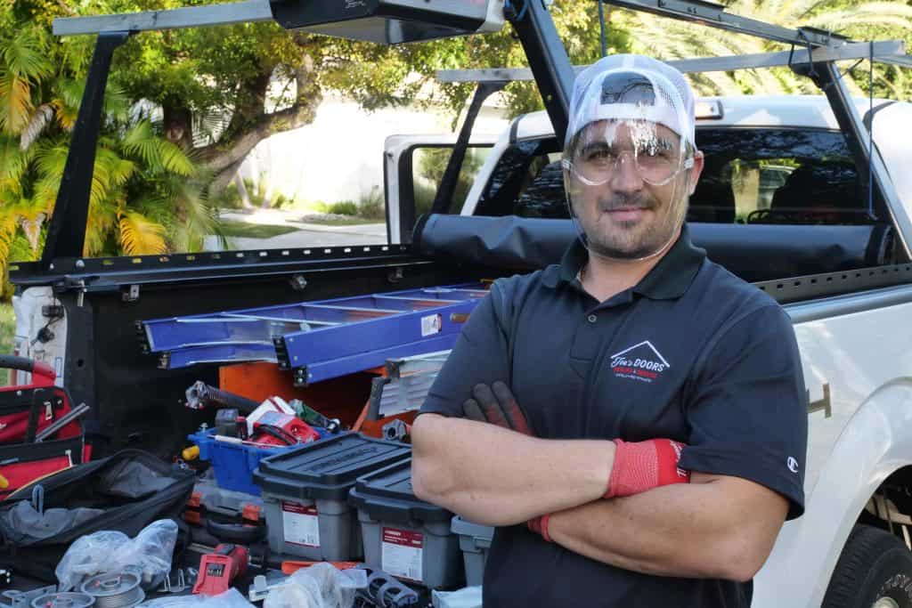 A man wearing a cap and safety glasses stands confidently with arms crossed next to a pickup truck loaded with tools and equipment. He wears a black shirt and gloves, and palm trees are visible in the background.