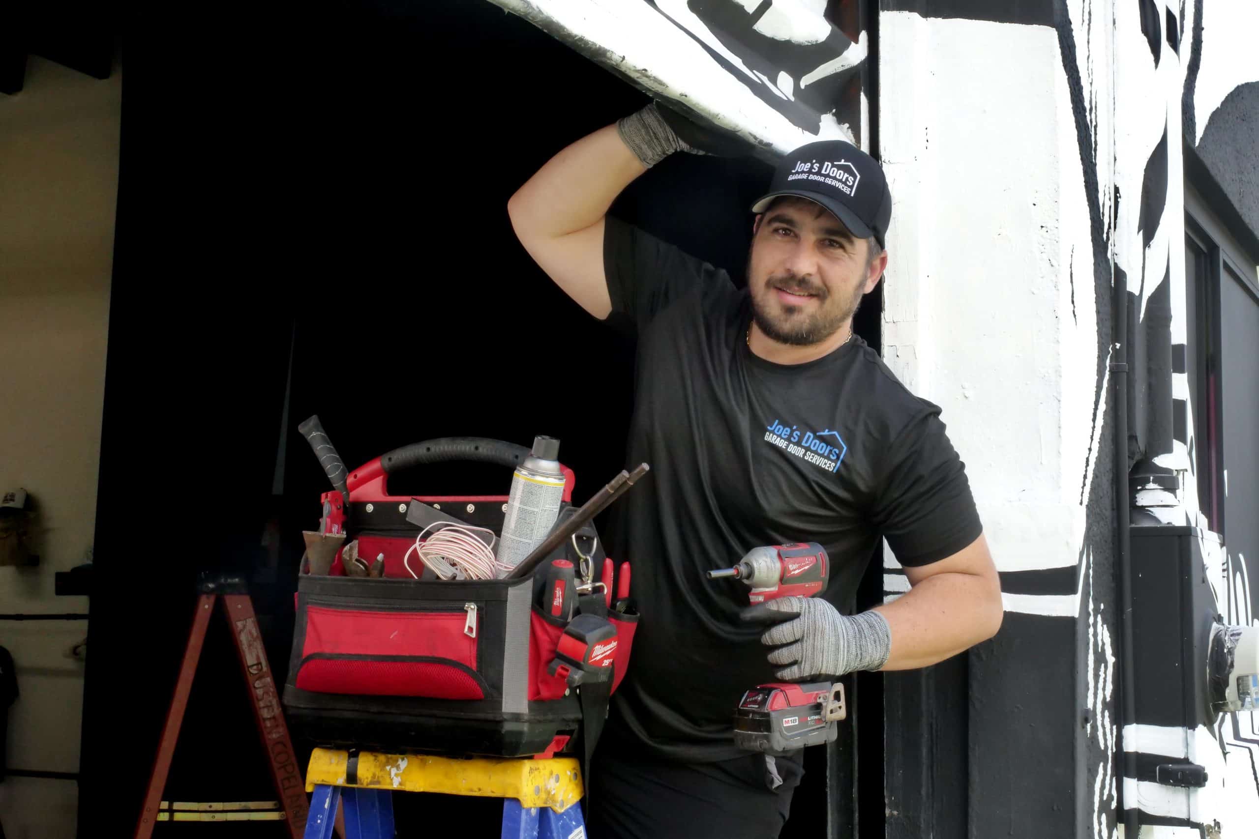 A man in a black t-shirt and cap stands on a ladder, holding a power drill and supporting a garage door with one hand. A red and black tool bag is next to him. He is in a workshop setting with a black and white mural in the background.