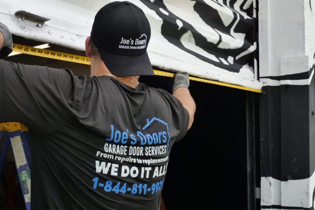 A worker in a "Joe's Doors Garage Door Services" shirt and cap measures a garage door with a tape measure. His shirt advertises repairs, replacements, and a phone number, showcasing professional garage door repair in HOG TOWN.