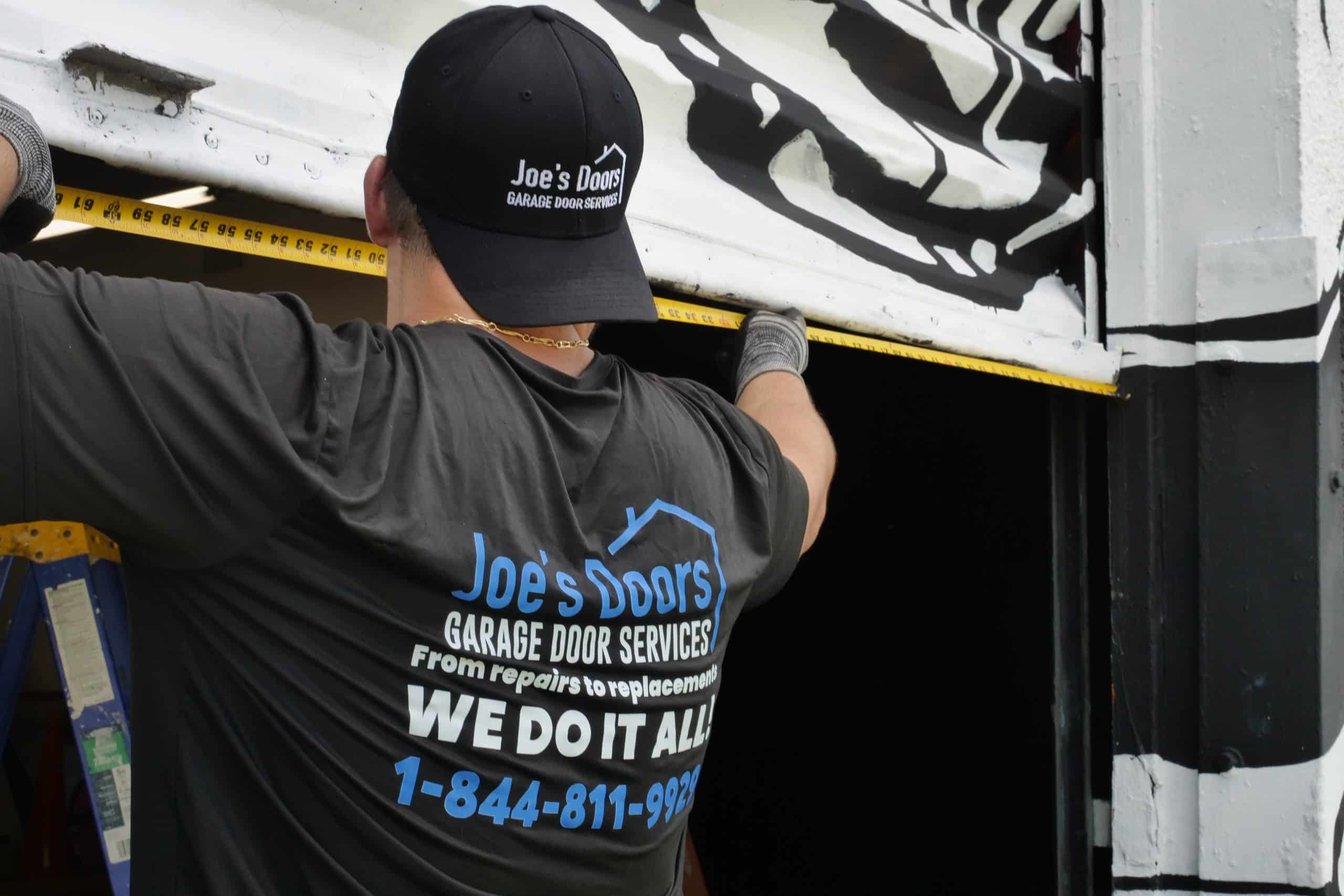 A worker in a "Joe's Doors Garage Door Services" shirt and cap measures a garage door with a tape measure. His shirt advertises repairs, replacements, and a phone number, showcasing professional garage door repair in HOG TOWN.