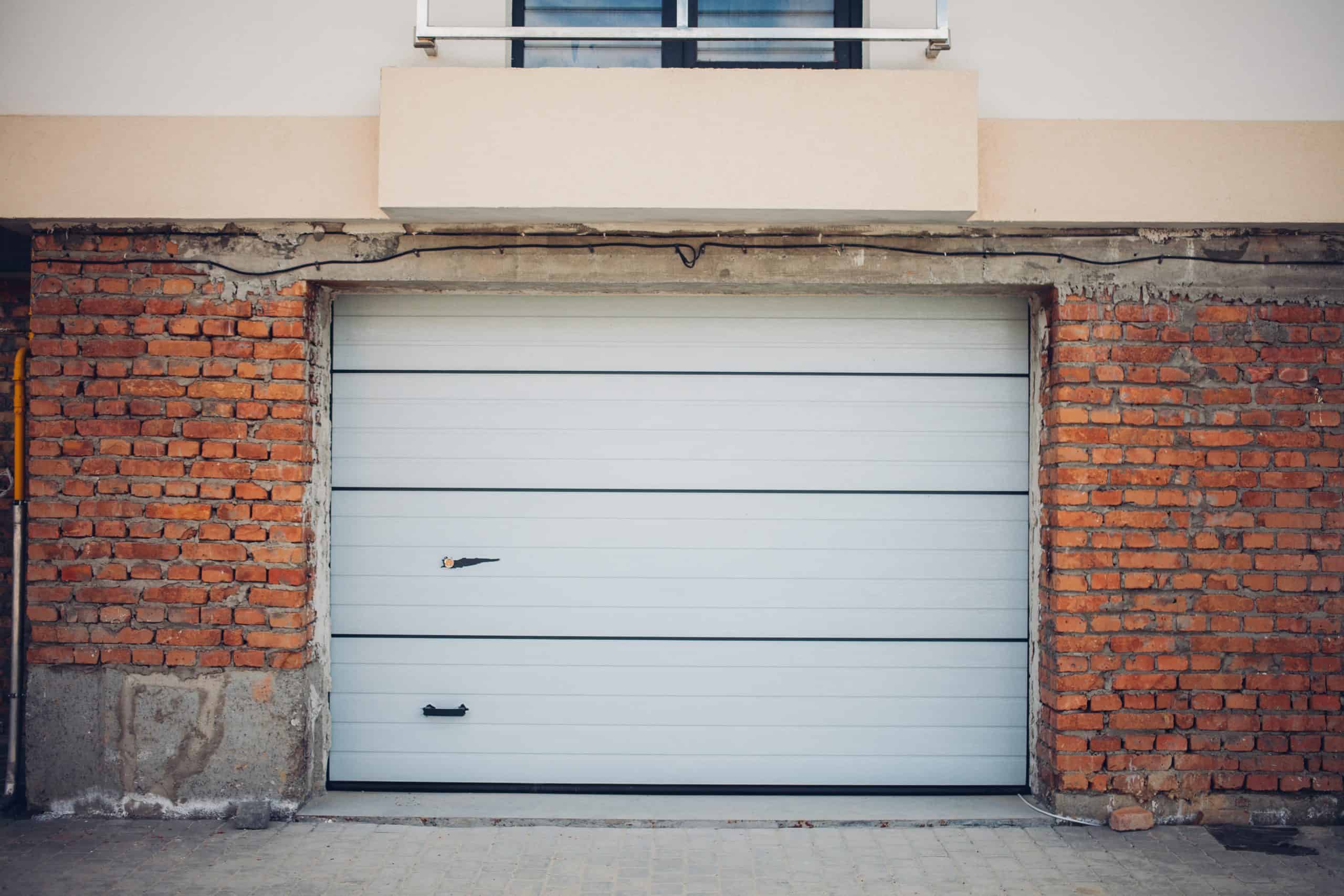 A closed white garage door set in a brick wall with a narrow upper window. The wall has areas of exposed brick and unfinished edges, giving a rustic appearance. The ground in front is paved with light-colored bricks.