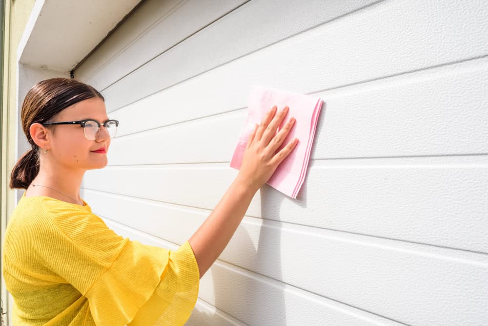 A person wearing glasses and a yellow shirt cleans a white garage door with a pink cloth. The image is taken during the day, and the person appears focused on the task.