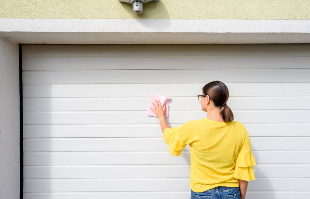 A person in a yellow sweater and jeans is cleaning a white garage door with a pink cloth. They have dark hair tied back, and wear glasses. The garage wall above the door is light green.
