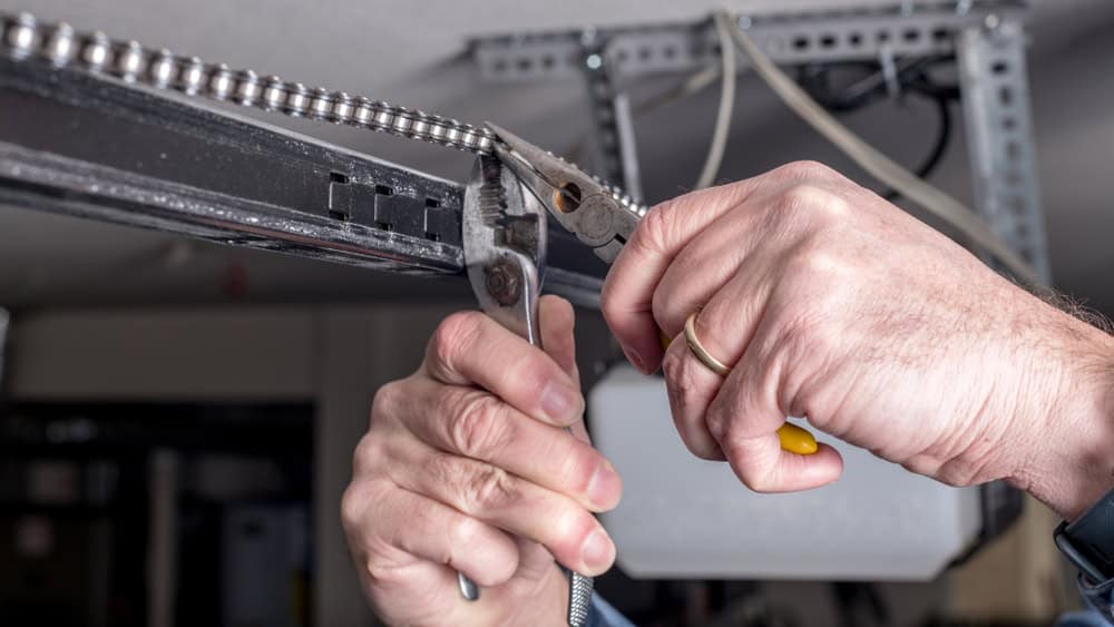 A person uses pliers to adjust a metal gear chain on a garage door mechanism. The person's hands are the focus of the image, with a light-colored background and metal framework visible.