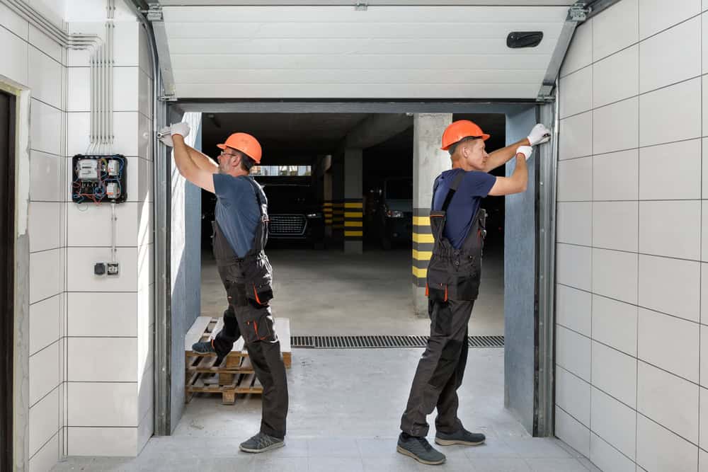 Two workers wearing orange helmets and dark overalls are installing a garage door. They are in a tiled area with tools and a partially open garage space behind them, where a car and striped safety poles are visible.