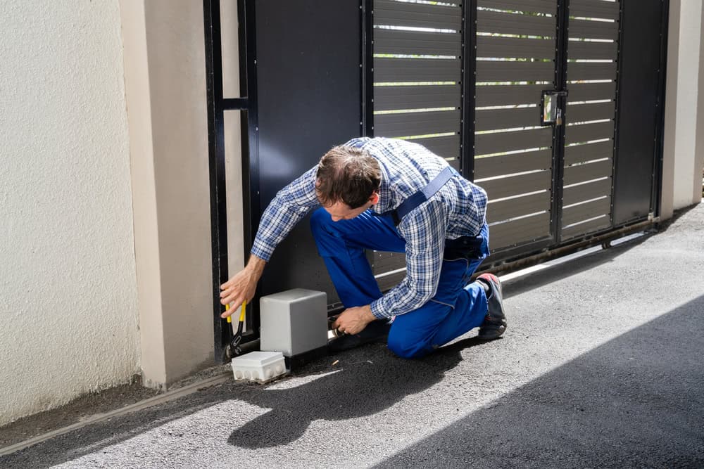 A man in a checkered shirt and blue overalls is kneeling on the pavement, installing or repairing a mechanism near a metal gate. The man is holding a tool and working on a rectangular box fixed at the base of the gate.