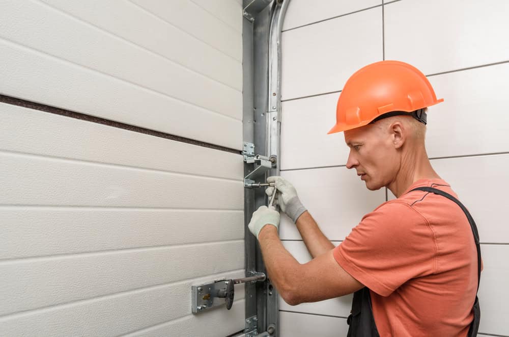 A construction worker in an orange hard hat and gloves tightens a bolt on a sectional garage door using a wrench. The worker is focused on adjusting the metal brackets on the door's vertical track.
