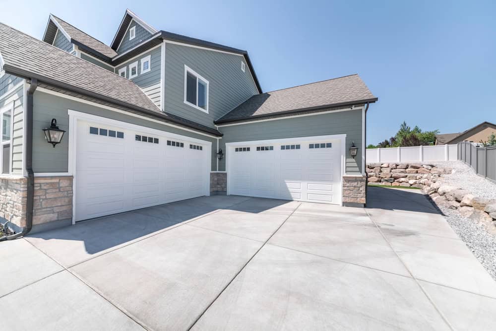 A large house with gray siding and a stone accent features three white garage doors. The driveway is expansive and concrete, and there's a landscaped area with rocks and shrubs on the right. The sky is clear and blue.