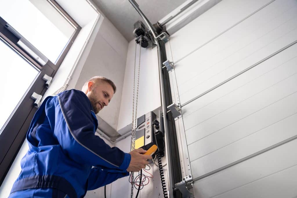 A technician in a blue uniform is working on a garage door opener system. He is adjusting or inspecting the control panel mounted on the wall next to the door. The setting appears to be a bright, industrial space.