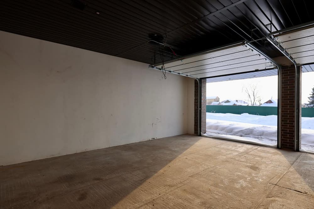 Empty garage with raised door, revealing a snow-covered driveway and fence outside. The interior features a smooth concrete floor, white walls, and a dark ceiling. Natural light from the open door illuminates the space.