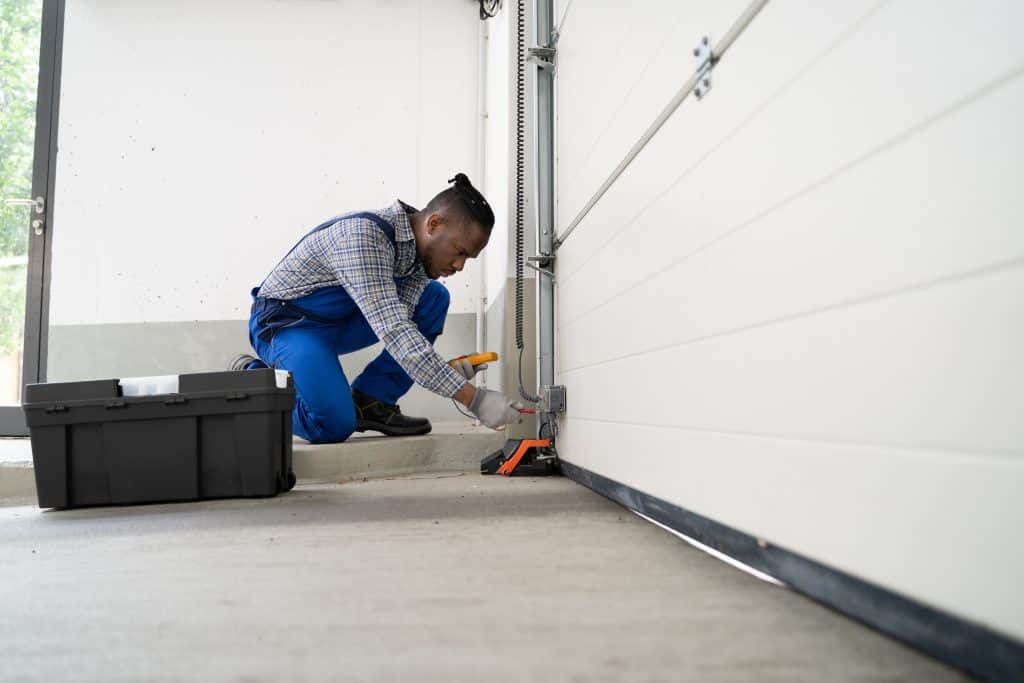 A person in blue overalls and a plaid shirt is kneeling and working on the bottom of a garage door. They have a toolbox nearby and appear focused on repairing or adjusting the mechanism.