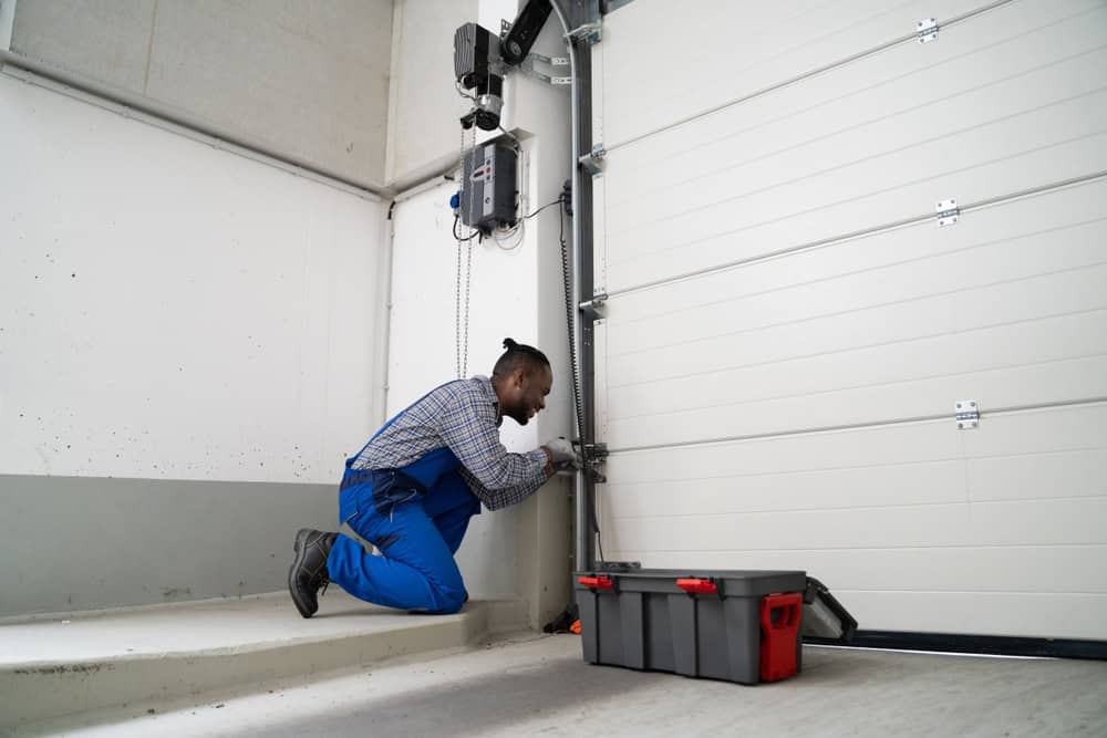 A person in blue overalls kneels on the floor, working on a garage door mechanism. A toolbox is open nearby. The setting appears to be a garage interior with white walls and a closed sectional garage door.