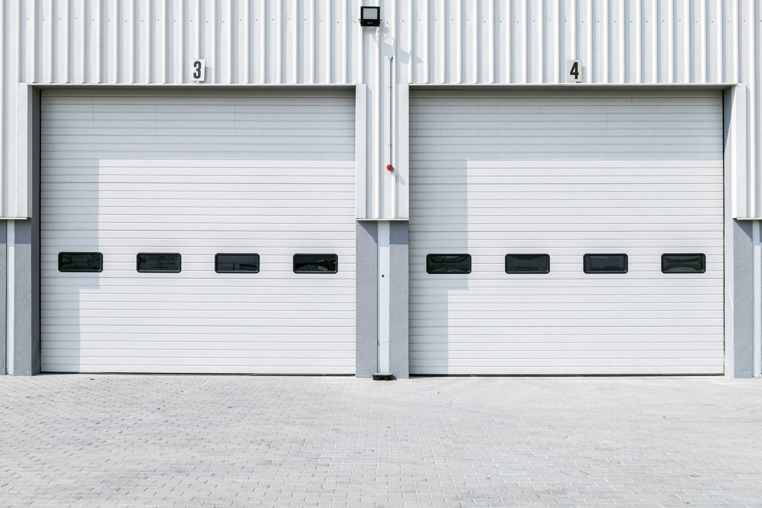 Two white industrial garage doors are side by side, labeled with numbers 3 and 4. Each door has a row of four rectangular windows. The ground in front is paved with light gray bricks.