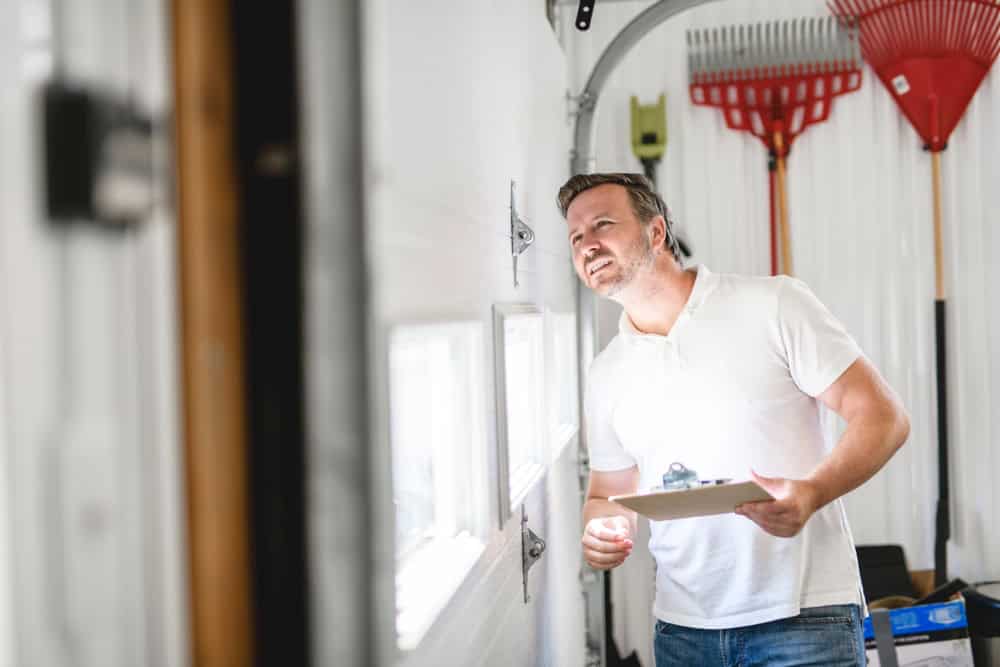 A person in a white polo shirt stands in a garage, holding a clipboard and inspecting a garage door. Several gardening tools hang on the wall in the background, including rakes.