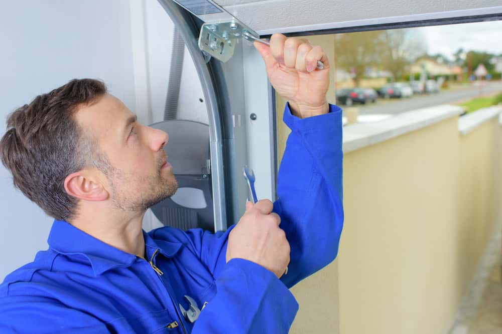 A man in a blue uniform is repairing a garage door. He is using a wrench to tighten a bolt on the upper edge of the door. He appears focused on his task with a residential street visible in the background.