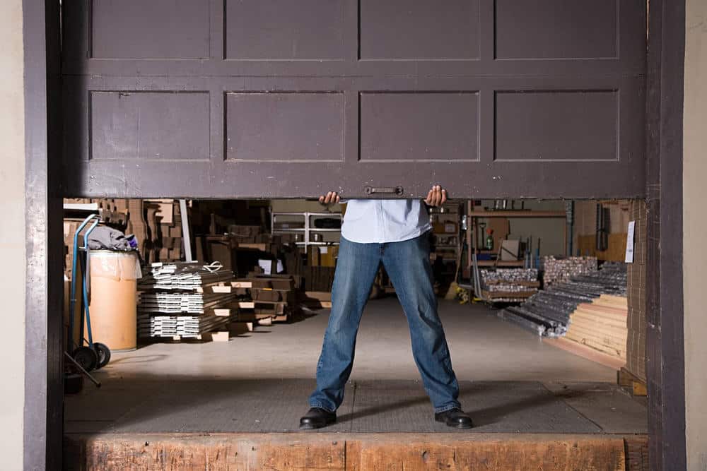 A person wearing jeans and black shoes lifts a large garage or warehouse door from the inside. The space is filled with various tools, stacks of wood, and a dolly with cardboard tubes.