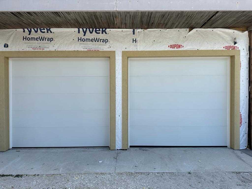 Two closed white garage doors stand side by side in a building under construction—Tyvek HomeWrap and Dupont insulation frame the unfinished sections beside the doors, part of Job 1836 overseen by Mallory Marshall.
