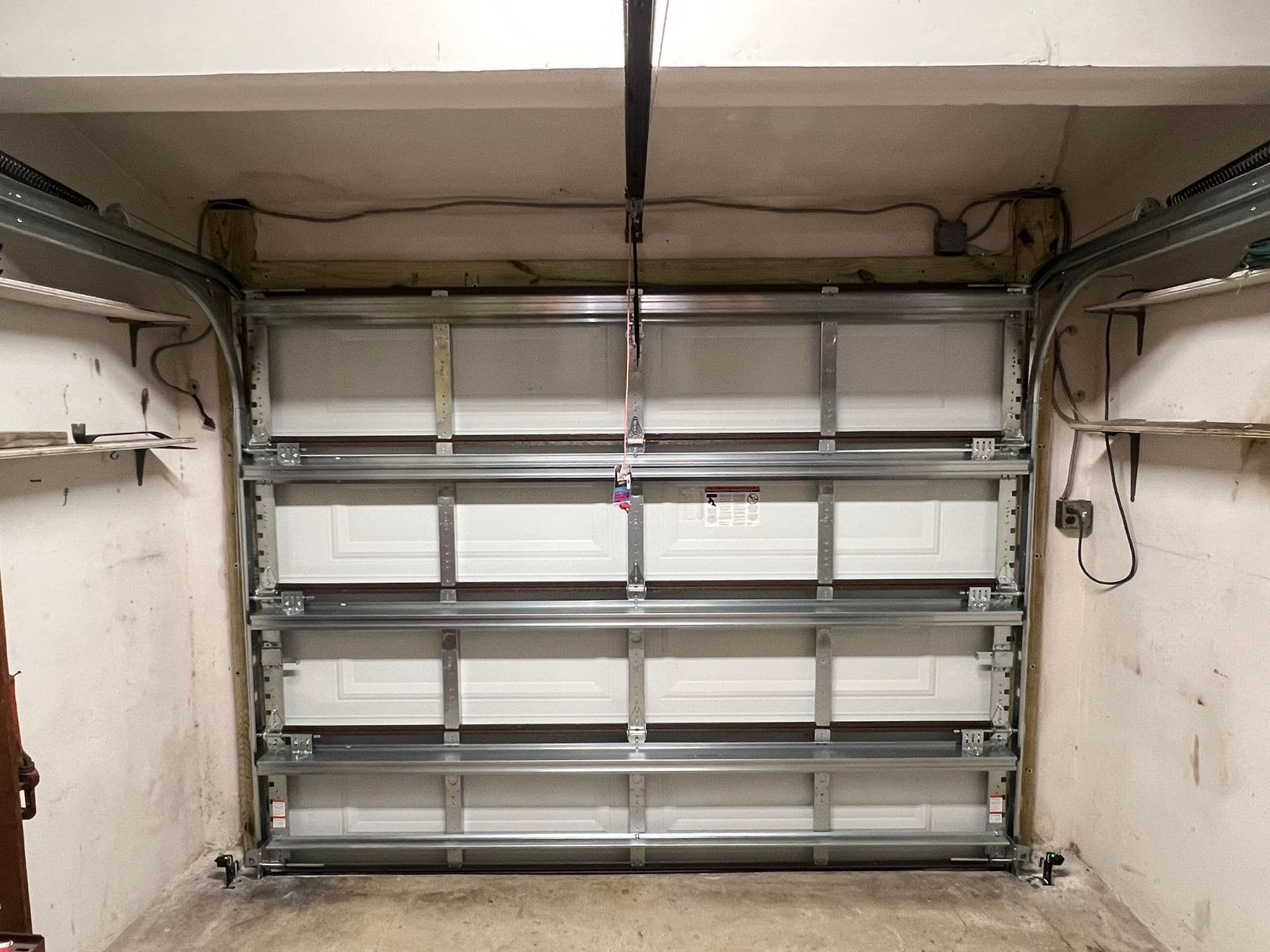 A closed white sectional garage door with metal supports inside a garage, surrounded by beige walls, exposed wiring, and two shelves on either side. Job 5048 was completed by Benjamin Flores.