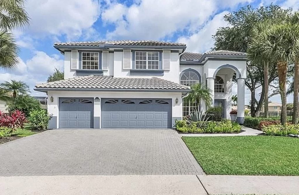 Two-story house with a tiled roof and three-car garage. The exterior is painted in white and gray tones. The front yard is landscaped with grass, bushes, and palm trees, set against a backdrop of a partly cloudy sky.