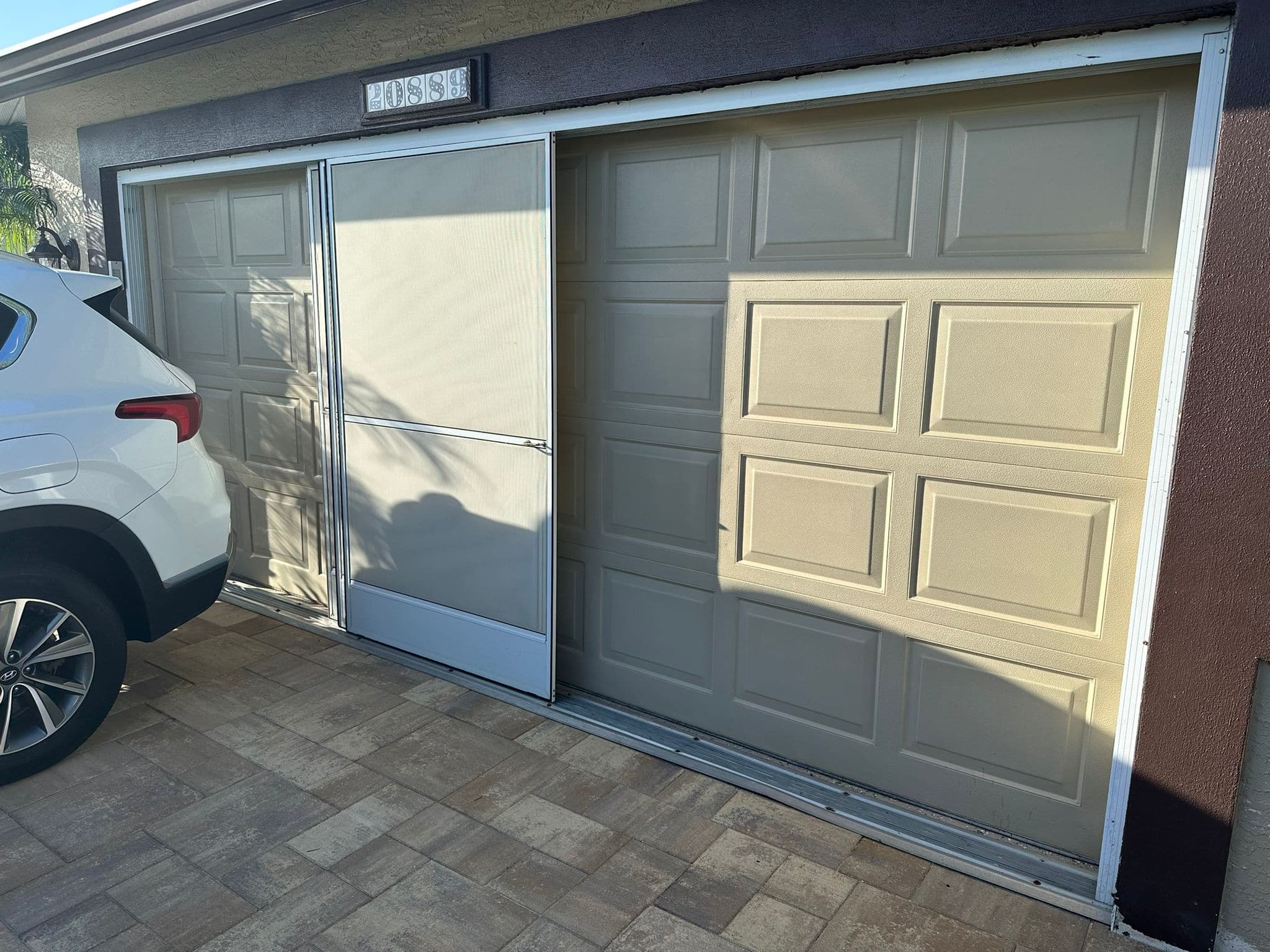 A white car is parked next to a garage with two beige doors and a screen door in the center. The area, part of Job 5146 by Richard Butrim Sr, is paved with light brown tiles, and sunlight casts shadows across the surface.