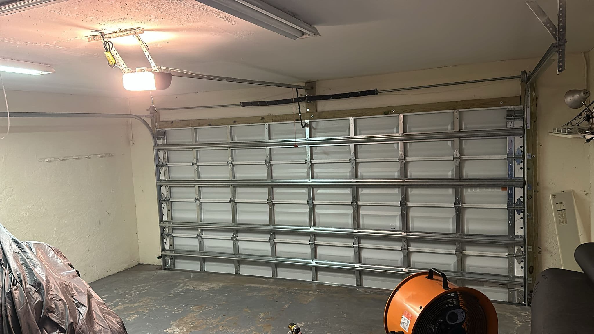 A closed, silver metal garage door inside a tidy garage with a concrete floor. An orange industrial fan sits on the ground, and part of a covered item is visible on the left. Overhead lights illuminate the space.
