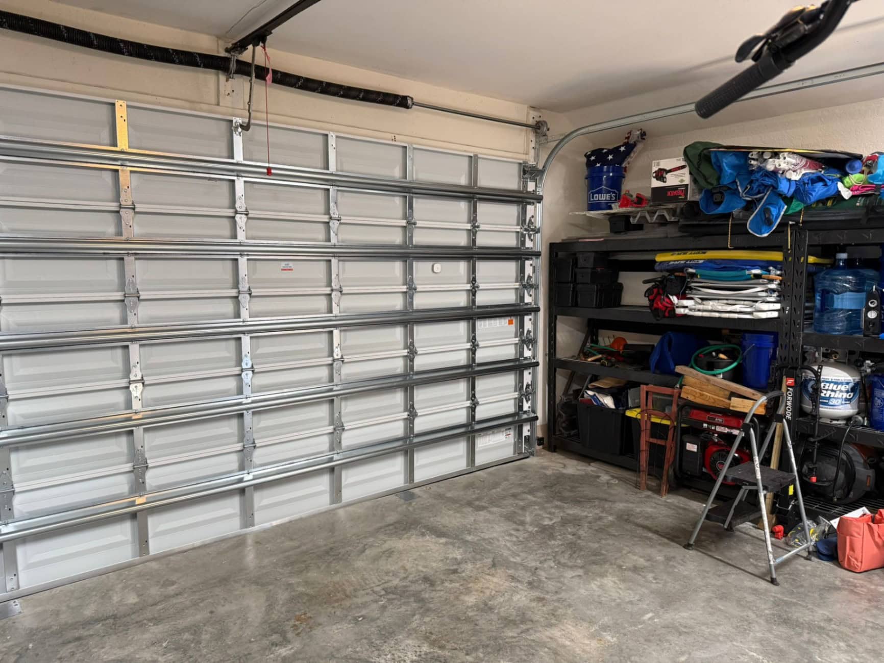 A clean garage with a closed metal door. On the right, organized shelves hold coolers, bins, water containers, and various tools from Job 5365. A folded step ladder sits in front of the shelves on a concrete floor by Daniel Royael.