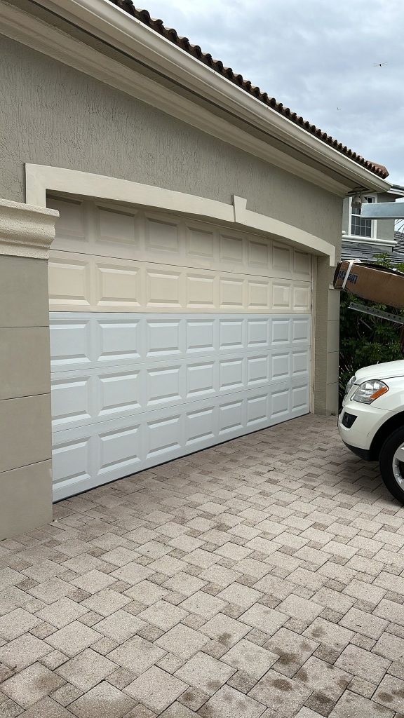 A garage door with mismatched panels—tan on the upper section, white on the rest—stands next to a white vehicle parked on a stone driveway. Photo for Job 4351 by Anna Kallenberg.