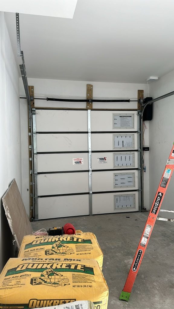 A residential garage with a closed sectional door, several control panels mounted on the door, a red and green ladder to the right, tools on the floor, and bags of Quikrete concrete mix in the foreground from Steve Saltouridis’s Job 4557.