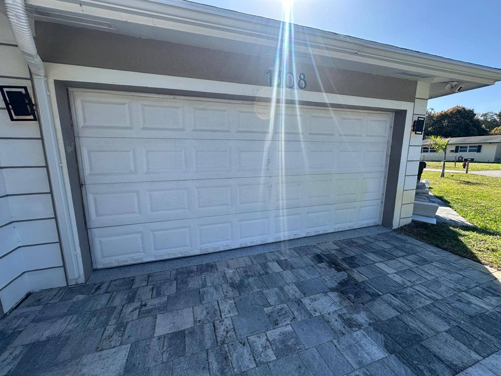 A white garage door with rectangular panels is closed, sunlight shining above it. The paved gray stone driveway sits at a Pompano Beach home in 33060, with a small green lawn and neighboring house visible to the right.