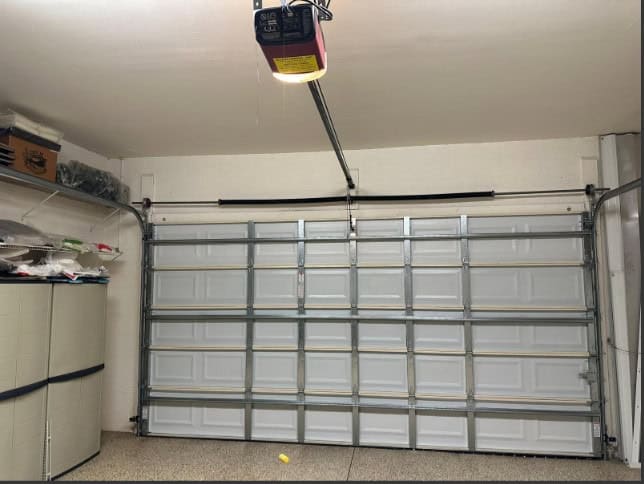 A clean, empty garage with a closed sectional garage door, shelves on the left wall storing various items, and a ceiling-mounted garage door opener. Job 1610 by Brad Heiges. A small yellow ball rests on the floor.
