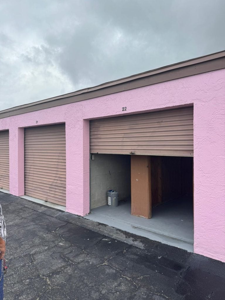 A row of storage units with pink walls and brown doors, one labeled 22 is partially open—revealing a trash can and wooden divider inside. The ground is paved, the sky overcast, and Job 4569 for Beverly Miller is underway.