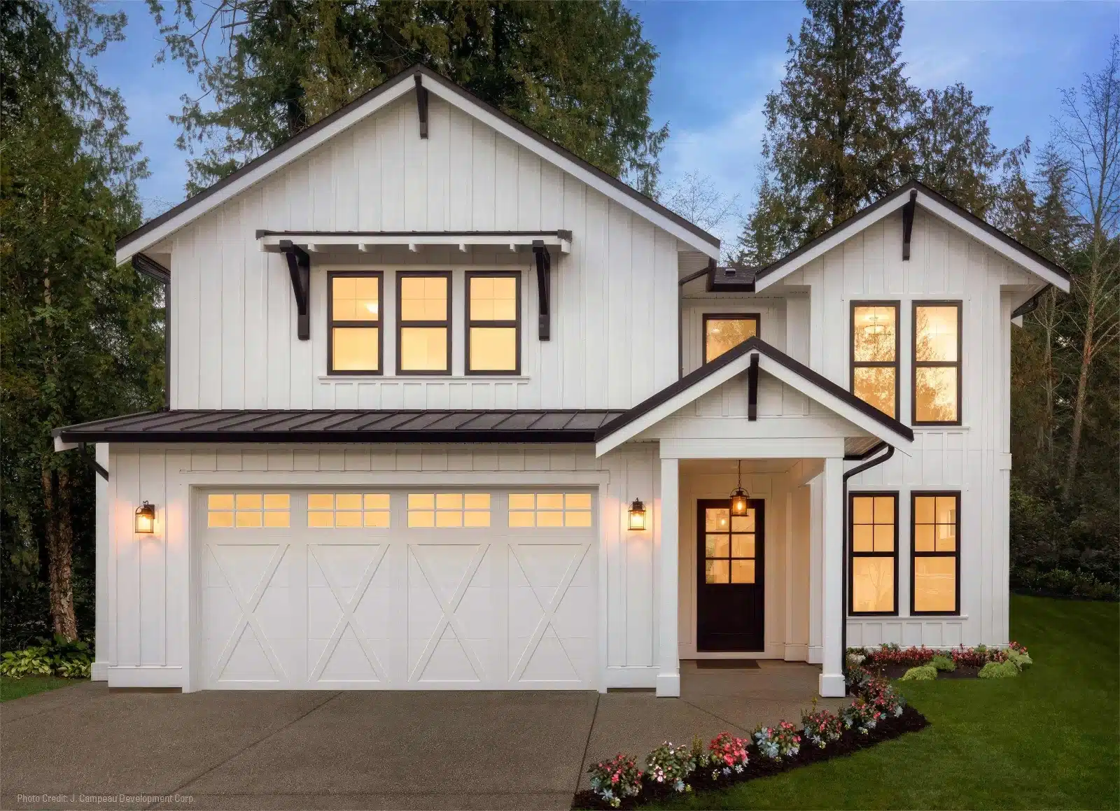 A modern two-story house with white siding, black trim, large windows, and a double garage. The entrance is well-lit, flower beds line the walkway, and Garage Door Repair ensures the double garage stays functional amid a lush lawn and tall trees.