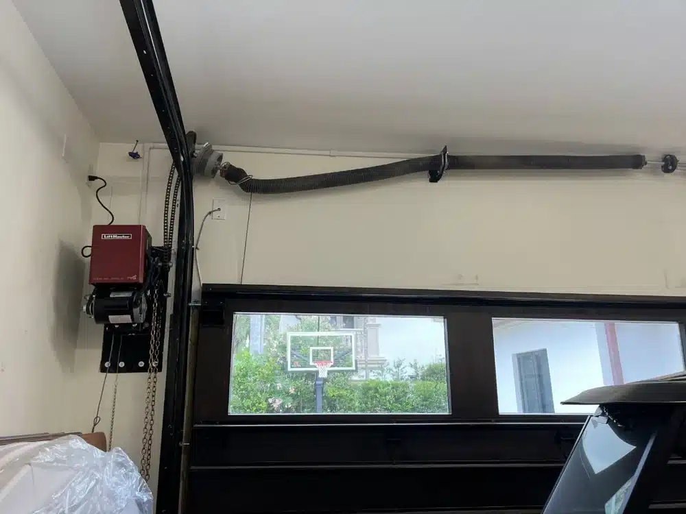 A garage interior showing an overhead door mechanism, an automatic garage door opener on the left wall, black metal tracks, and a basketball hoop visible outside through the windows—ideal for showcasing Garage Door Repair services.