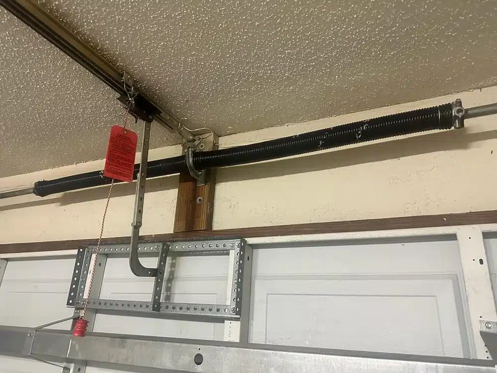 A close-up view of a garage door mechanism shows a black torsion spring, metal supports, and a red emergency release handle hanging from the opener track—highlighting details often inspected during Garage Door Repair services. The ceiling has a textured finish.