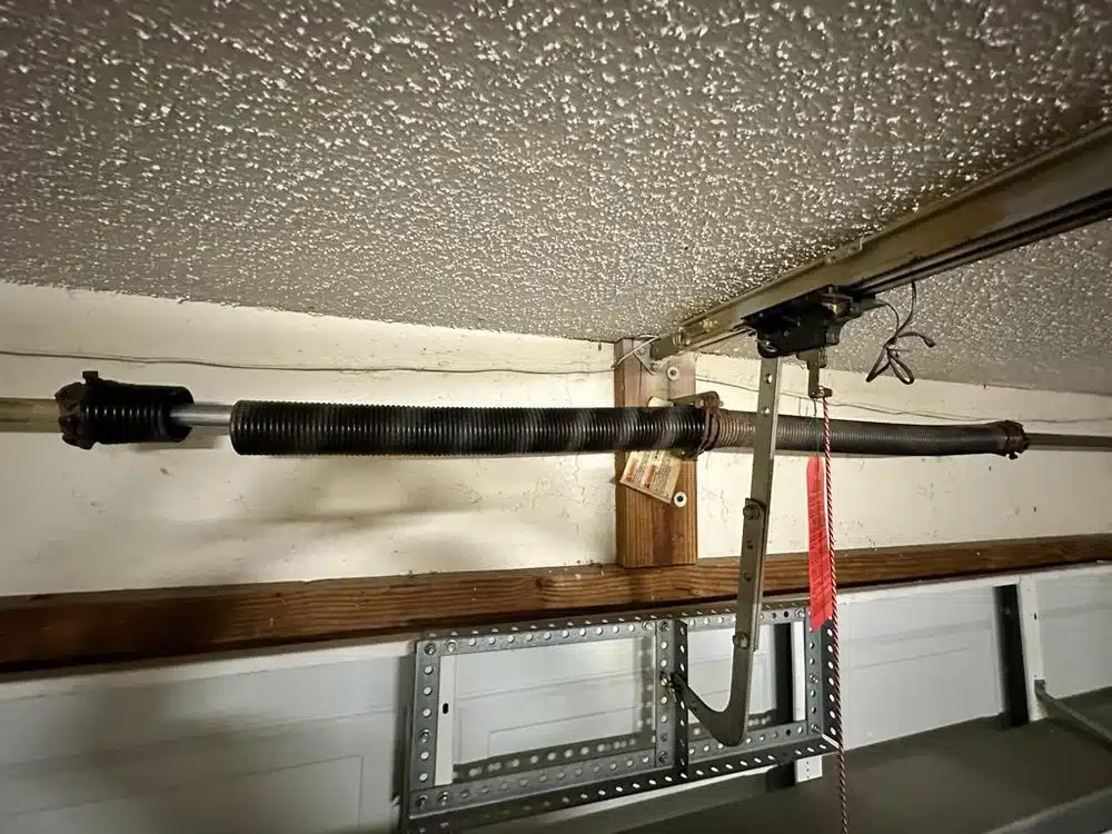 A close-up of a broken garage door torsion spring mounted on a metal rod above a garage door, with part of the garage door opener and a red pull cord visible—highlighting the need for professional Garage Door Repair services.
