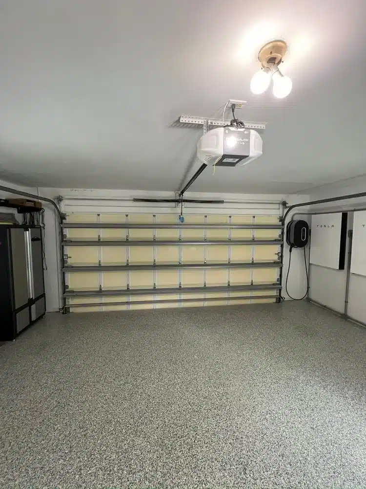A clean, empty garage with speckled flooring, a closed metal garage door, overhead lighting, a Tesla wall charger mounted on the right wall, and shelves and a refrigerator on the left—perfect for showcasing reliable Garage Door Repair services.
