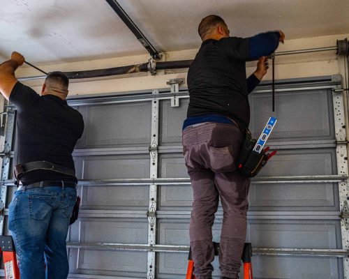Two men standing on ladders, working together on a HOG TOWN Garage Door Repair, fixing or installing a metal garage door mechanism in a residential garage.