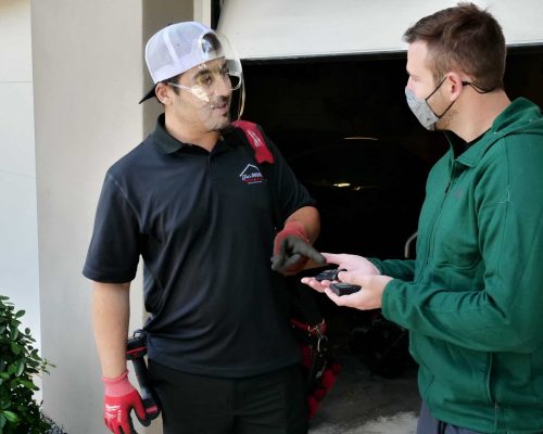 A worker in a black shirt, gloves, and face shield talks to a man in a green jacket and gray face mask holding a car key fob outside a HOG TOWN Garage Door Services garage.
