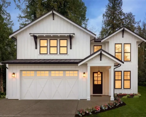 A modern two-story house with white siding, black trim, large windows, and a double garage. The entrance is well-lit, flower beds line the walkway, and Garage Door Repair ensures the double garage stays functional amid a lush lawn and tall trees.