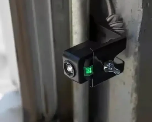 Close-up of a small black security camera with a glowing green indicator light, mounted on a metal surface near tools commonly used for garage door repair services; a wingnut and cable are visible behind it.